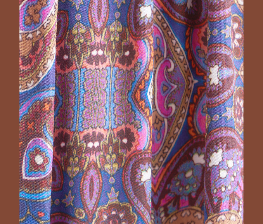 Nahaufnahme eines lebhaften Stoffes mit einem aufwendigen Paisley-Muster in Blau-, Lila-, Pink-, Orange- und Brauntönen mit weißen Akzenten, der vertikale Falten zeigt.