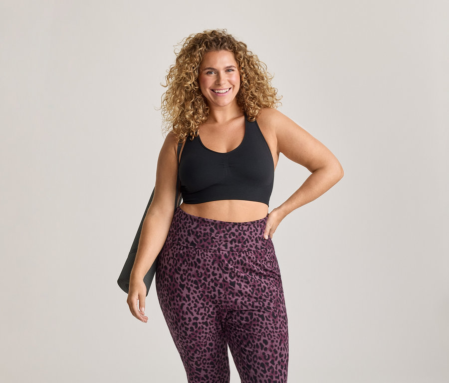 Lächelnde Frau mit lockigem Haar trägt einen schwarzen Sport-BH und lila Leggings mit Leopardenmuster, eine Yogamatte über der Schulter haltend.