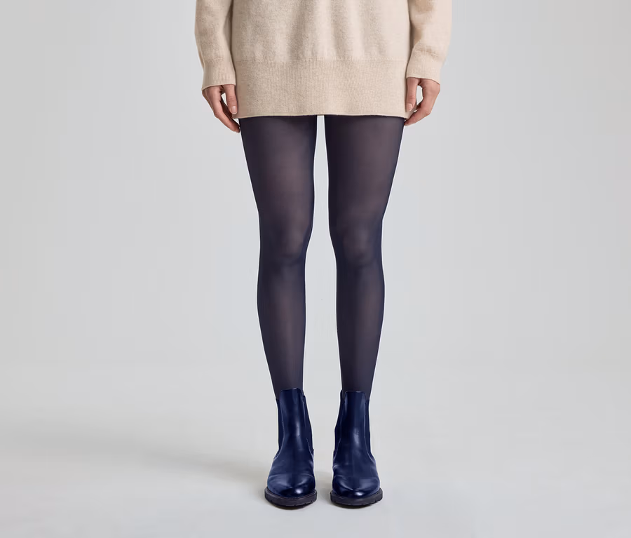 Person steht, trägt einen beigen Pullover, dunkelblaue Strumpfhosen und dunkelblaue Stiefeletten.