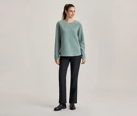 Stehende Frau in einem hellgrünen, strukturierten Pullover, schwarzen Hosen und schwarzen Schuhen, mit Pferdeschwanz.