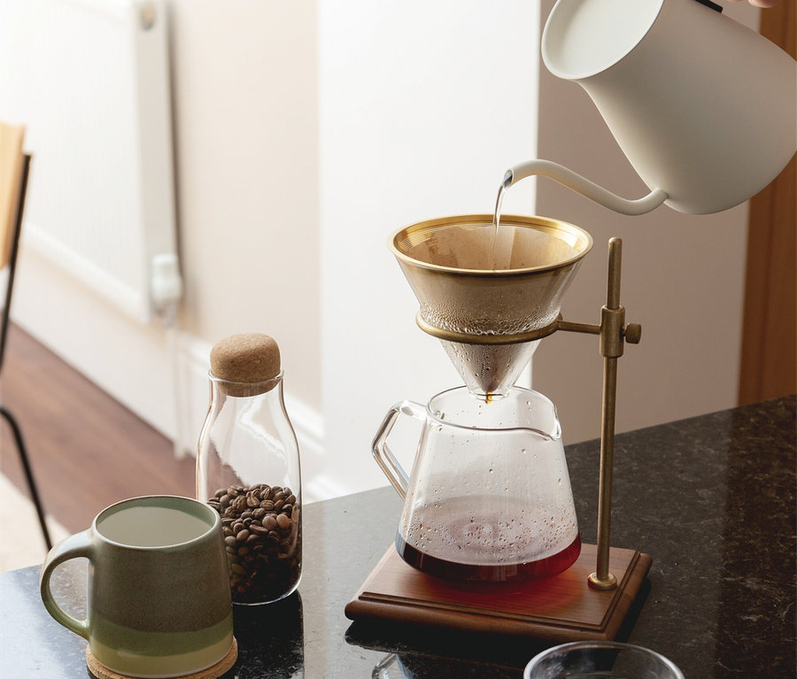 Eine Hand gießt Wasser aus einem weißen Schwanenhalskessel in einen goldenen Pour-over-Kaffeefilter, der auf einem Messingständer über einer Glaskaraffe steht. Auf der dunklen Arbeitsplatte befinden sich eine grüne Tasse, ein Glas mit Kaffeebohnen und ein Wasserglas.