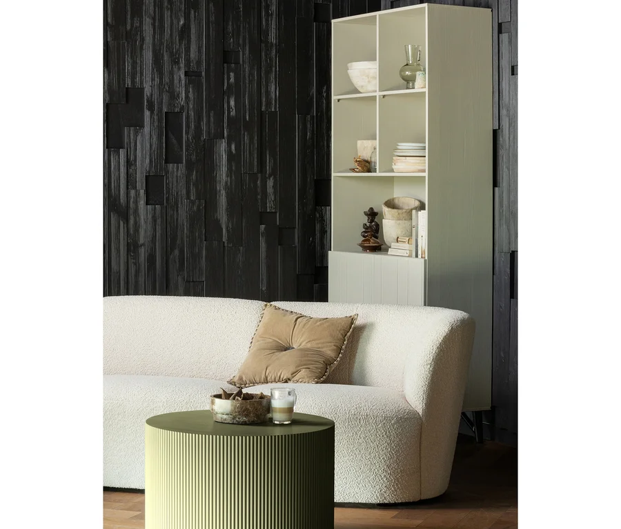 Ein cremefarbenes Bouclé-Sofa mit braunem Kissen steht vor einer dunklen Holzwand, neben einem hellen Regal. Ein runder, olivgrüner geriffelter Couchtisch trägt eine Dekoschale und ein Glasgetränk.