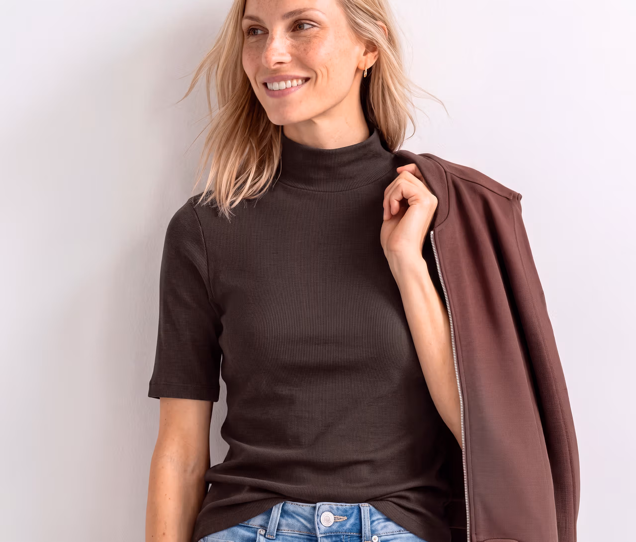 Lächelnde Frau mit blondem Haar trägt ein braunes geripptes Street One Shirt mit Rollkragen und kurzen Ärmeln, eine braune Jacke über der Schulter haltend.