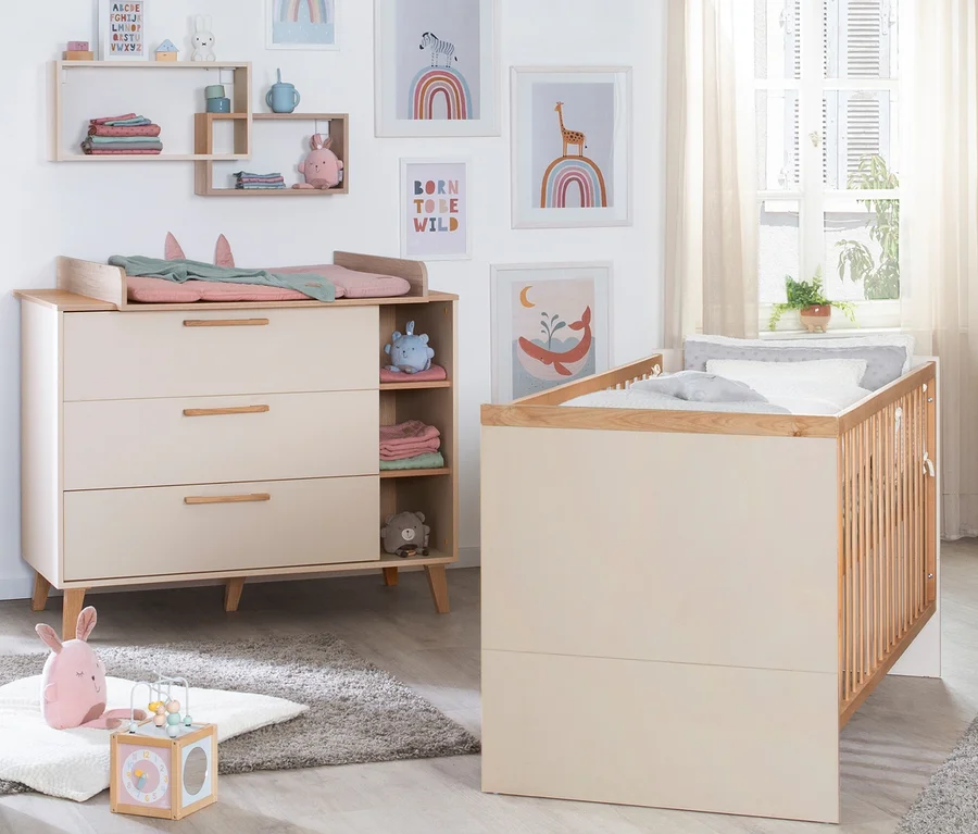 Ein neutral gehaltenes Babyzimmer mit einer hellbeigen Wickelkommode mit rosa Wickelauflage und einem passenden Holzbett mit weißer Bettwäsche. Wandregale zeigen gefaltete Kleidung und Spielzeug, während gerahmte Tier- und Regenbogenbilder die Wand schmücken. Ein grauer Teppich und Spielzeug liegen auf dem Boden.