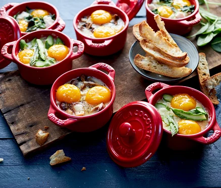 Mehrere kirschrote STAUB Keramik-Cocottes mit gebackenen Eiern, Pilzen, Spargel und Spinat, serviert mit geröstetem Brot auf einem Holzbrett.