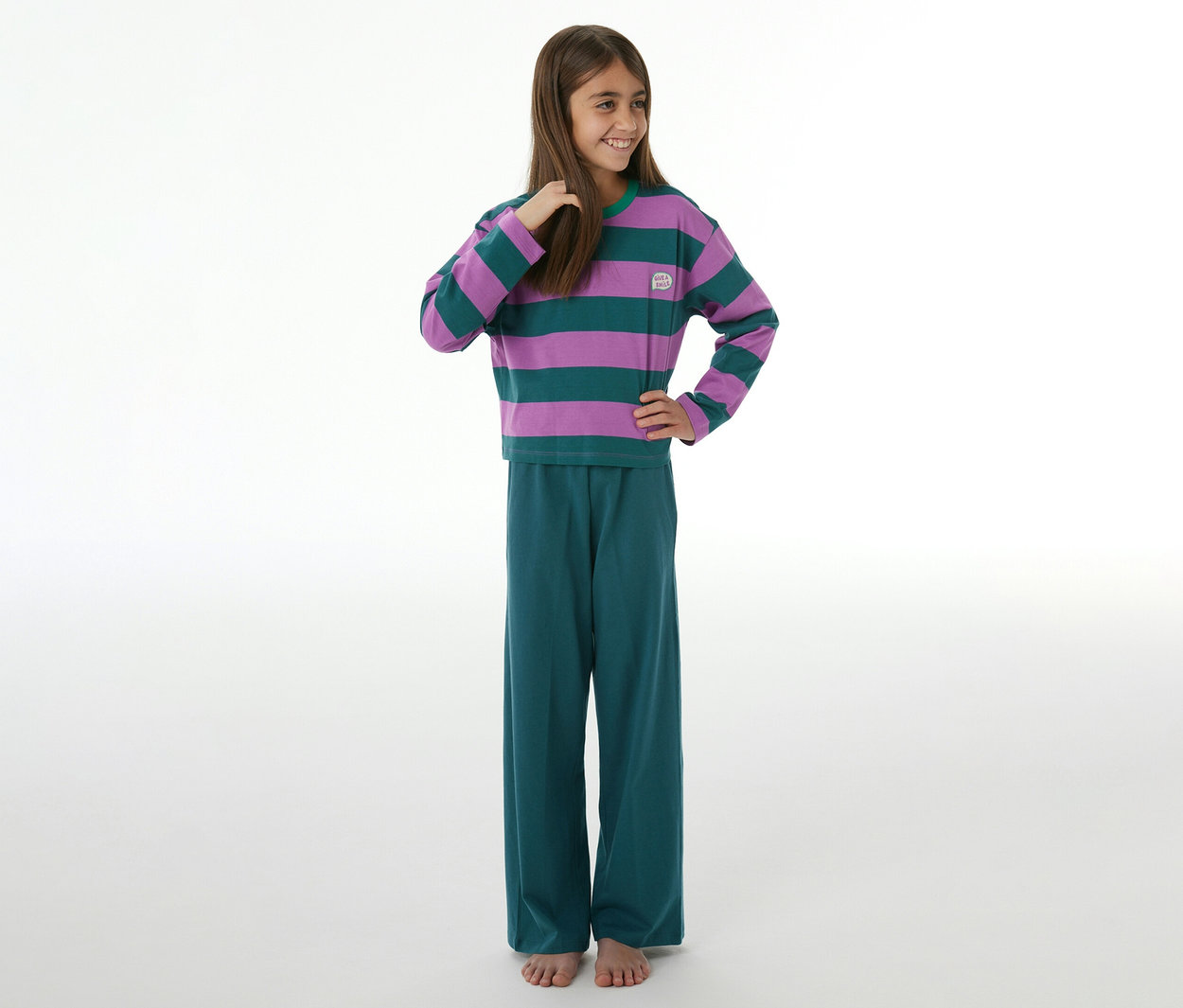 Lächelndes Mädchen in einem SCHIESSER Kinder-Pyjama mit lila-grün gestreiftem Langarmshirt und petrolfarbener Hose, barfuß.