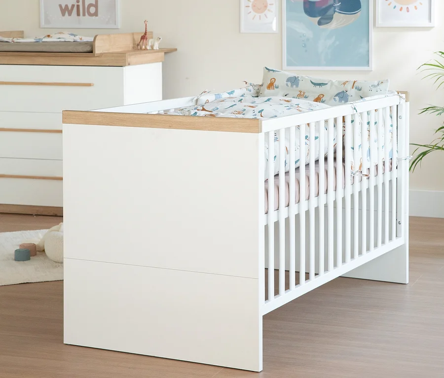 Ein weißes Babybett mit Holzrahmen und bunter Tierbettwäsche steht in einem Kinderzimmer neben einer passenden Wickelkommode.