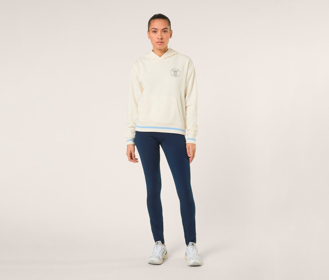 Frau steht, trägt einen cremefarbenen FILA Damen-Hoodie »MARLIA« mit hellblauen Streifen an Bündchen und Saum, dunkelblaue Leggings und weiße Sneaker.