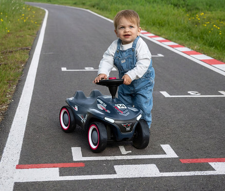 Kleinkind in Latzhose steht auf einer Rennstrecke und hält sich an einem dunkelgrauen BIG Bobby Car Neo RS mit rot-weißen Akzenten fest.