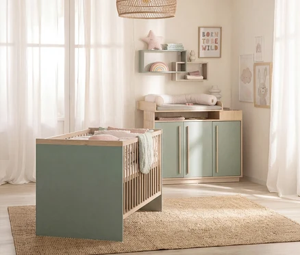 Babyzimmer mit einem hellgrünen Gitterbett und einer passenden Wickelkommode aus hellem Holz, auf einem beigen Teppich. An der Wand hängen Bilder und Regale.