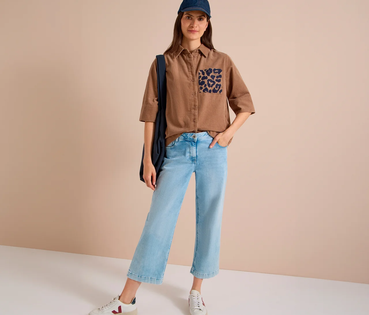 Stehende Frau in einer braunen CECIL Hemdbluse mit Leopardenmuster-Tasche, hellblauen Cropped-Jeans, blauer Baseballkappe und weißen Sneakern, eine dunkelblaue Tasche über der Schulter tragend.