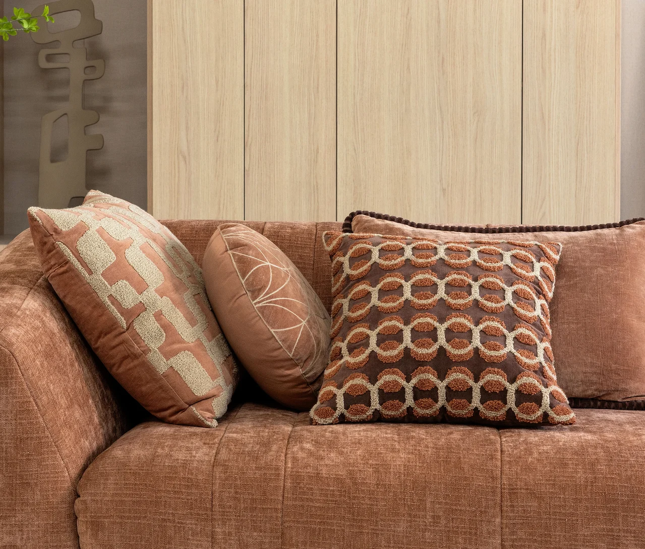 Vier WOOOD Dekokissen "Luvo Velvet" in verschiedenen Braun- und Beigetönen, mit unterschiedlichen Formen und Mustern, auf einem terrakottafarbenen Sofa.