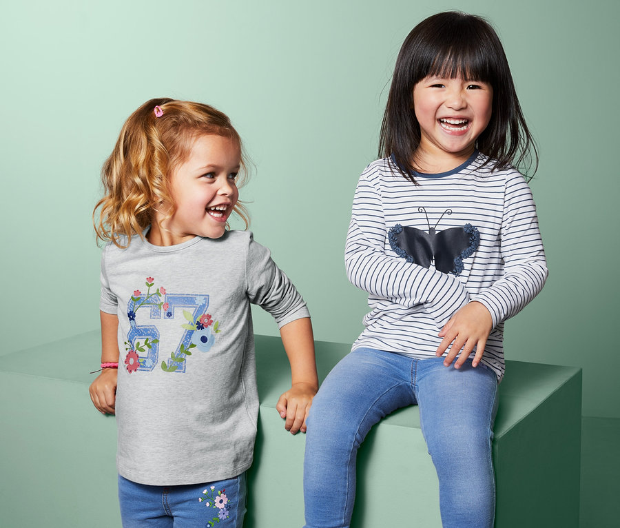 Zwei kleine Mädchen lachen und posieren. Ein Mädchen trägt ein graues Langarmshirt und Jeans, das andere ein gestreiftes Langarmshirt und Jeans.
