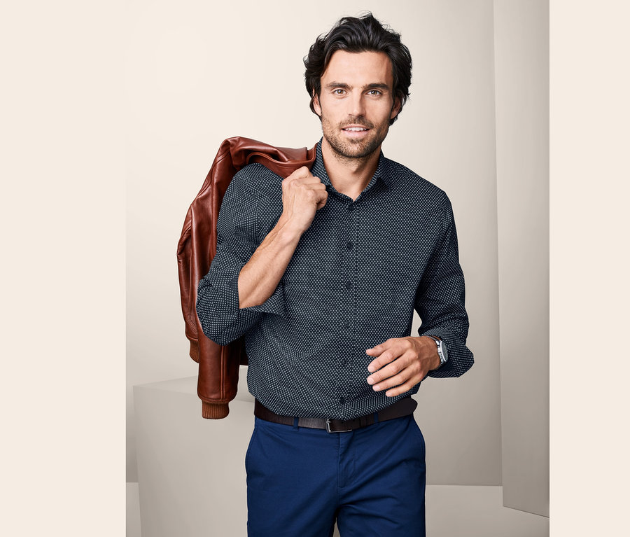 Mann mit brauner Lederjacke über der Schulter und blauem Hemd, Modern Fit.