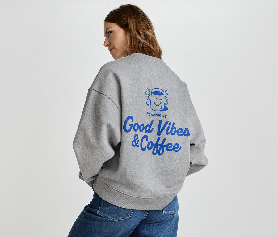 Frau trägt graues Sweatshirt mit blauem Aufdruck "Good Vibes & Coffee" auf dem Rücken, dazu Jeans.