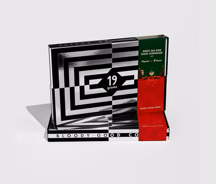 Teilweise geöffneter 19grams Adventskalender für Kaffee mit 24x 50 g ganzen Bohnen. Die Box ist schwarz-weiß gestreift, rot und grün, mit '19grams x Tchibo'.