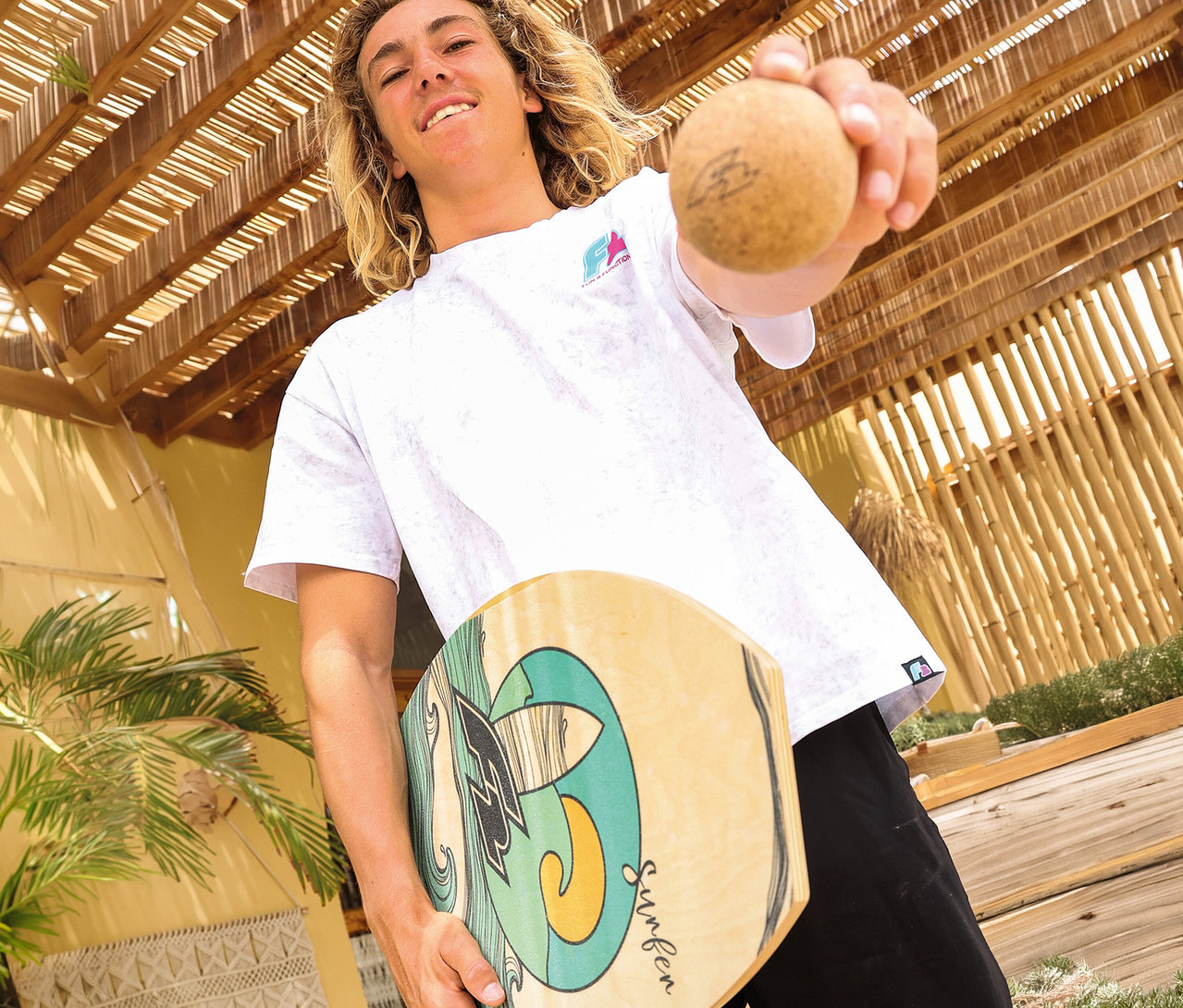 Ein lächelnder junger Mann mit langen blonden Haaren hält ein F2 Balance Board Drop mit farbigem Design und einen kleinen braunen Ball in die Kamera.
