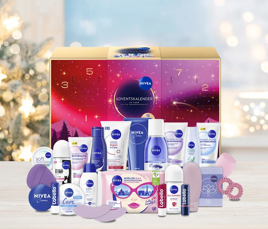 Ein goldener NIVEA Adventskalender mit lila-rosa Design und den Zahlen 1 bis 24 steht im Mittelpunkt, umgeben von einer Vielzahl von NIVEA Pflegeprodukten wie Cremes, Deodorants, Mizellen-Reinigungstüchern und Labello Lippenbalsamen. Im unscharfen Hintergrund sind festliche Lichter eines Weihnachtsbaums zu sehen.