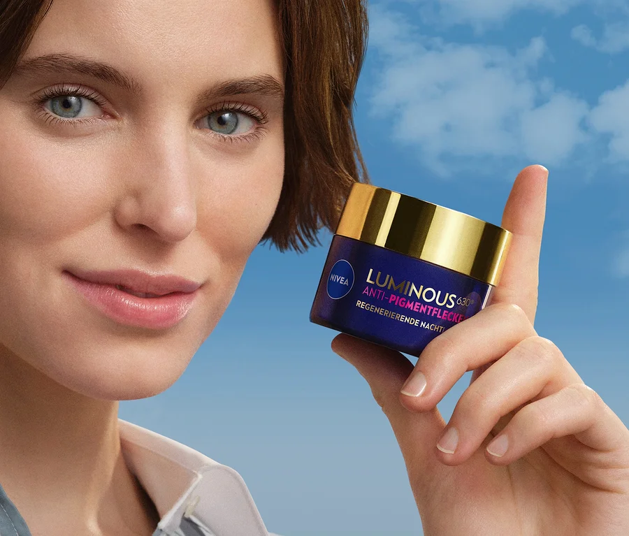 Lächelnde Frau mit kurzen braunen Haaren und blauen Augen hält einen dunkelblauen Nivea Luminous 630 Anti-Pigmentflecke Regenerierende Nacht Tiegel mit goldenem Deckel vor blauem Himmel.