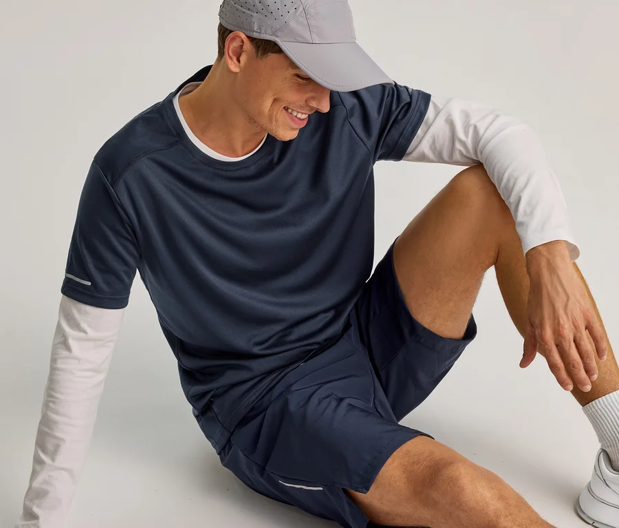 Lächelnder Mann sitzt auf dem Boden, trägt navy Sportshorts, ein navy T-Shirt über weißem Langarmshirt, graue Kappe und weiße Sneaker.