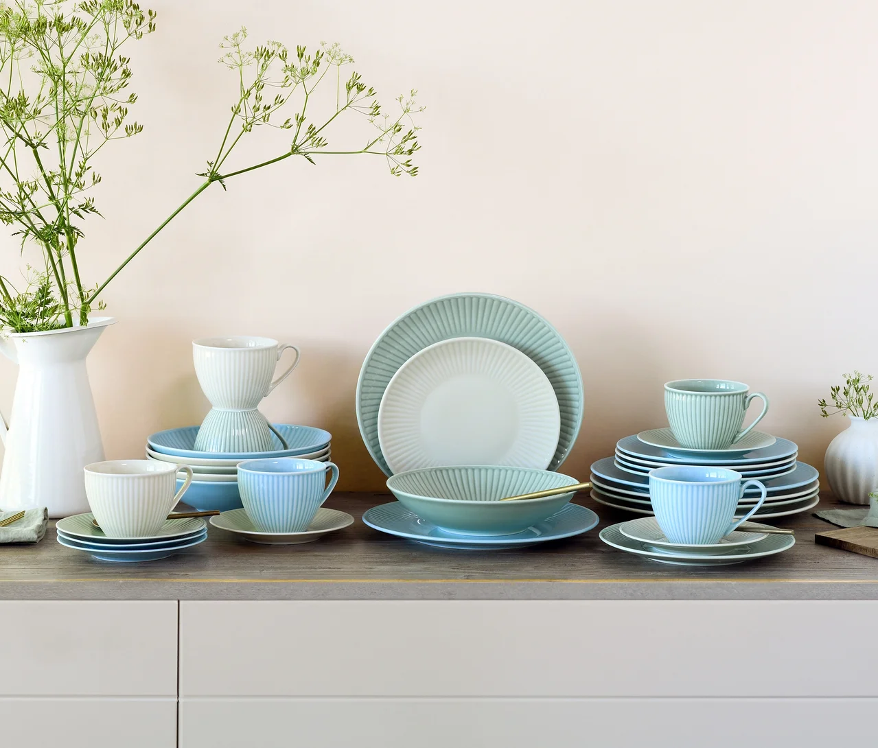 Ein creaTable Kombiservice »Valencia Pastell« mit Tellern, Schalen, Tassen und Untertassen in Pastellfarben wie Hellblau, Mintgrün und Off-White mit gerippter Textur, auf einer Holztheke arrangiert. Links steht ein weißer Krug mit grünen Pflanzen.