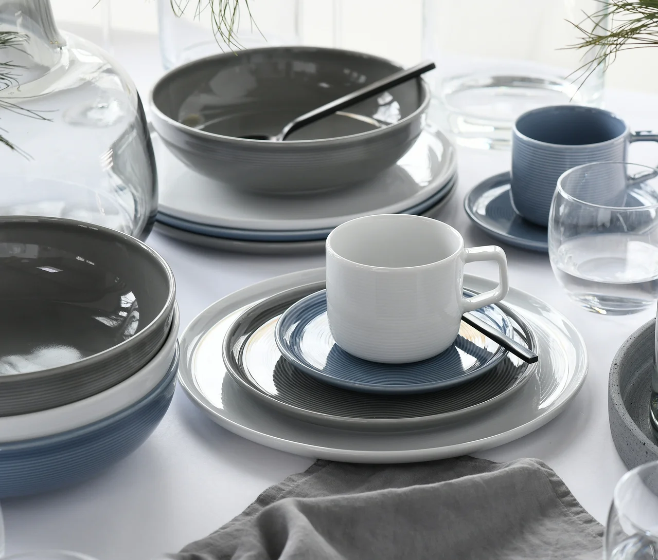 Ein Tisch gedeckt mit modernem creaTable Service »Session Urban«, 30-teilig, in Weiß, Grau und Blau, mit gerippten Tellern, Schalen und Tassen, dazu klare Glaskaraffen und Trinkgläser sowie eine graue Leinen-Serviette.