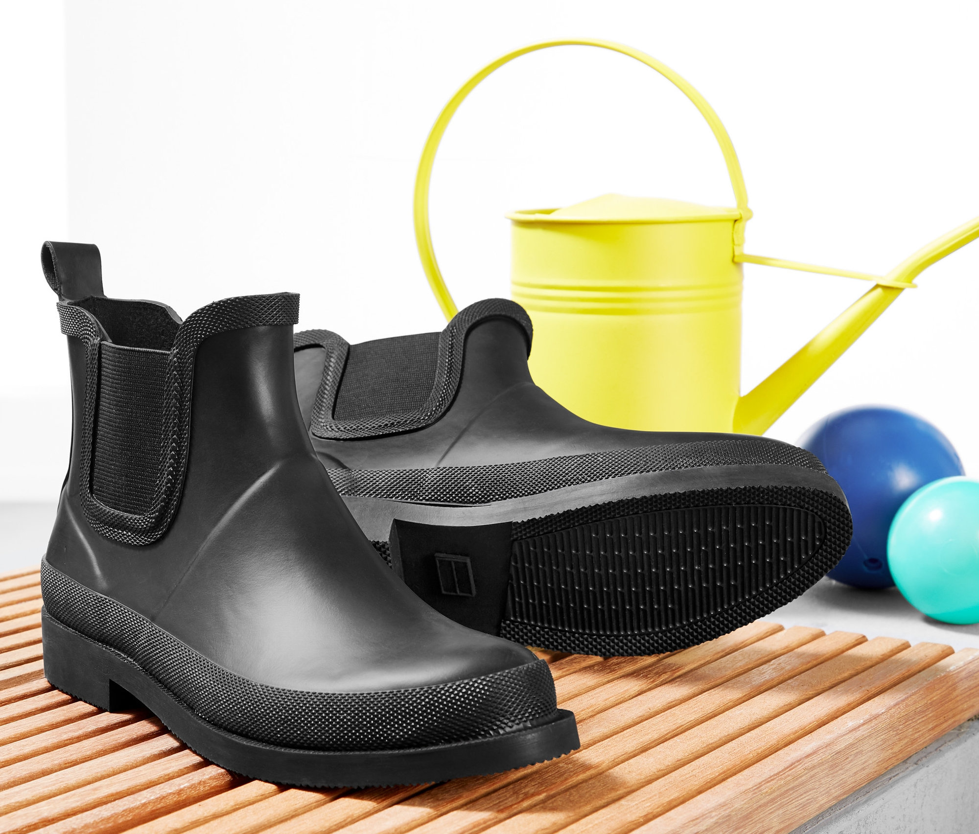 Zwei Paar schwarze Regenstiefeletten auf einer Holzbank, dahinter eine gelbe Gießkanne und zwei Kugeln.