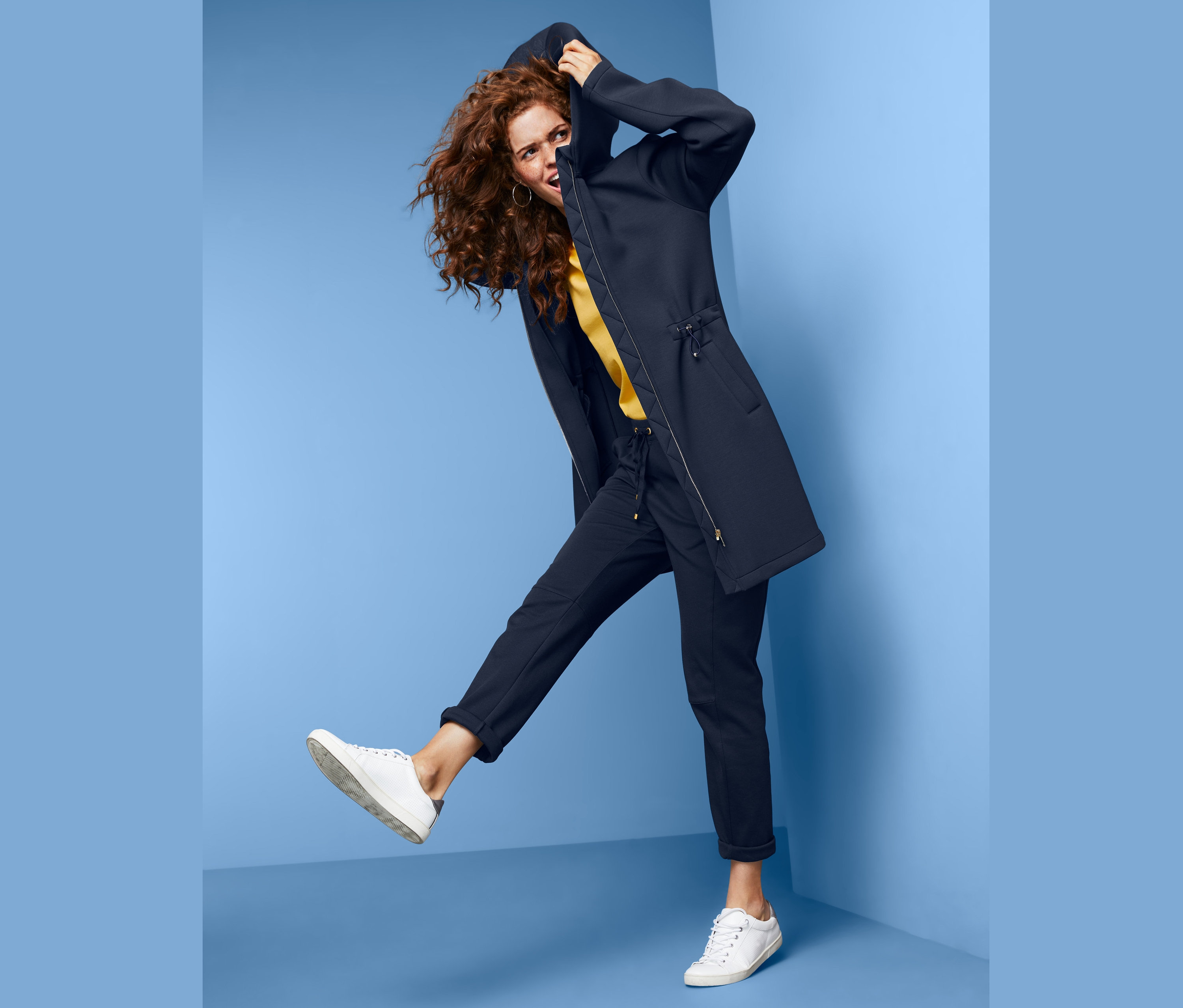 Frau mit lockigem Haar posiert in Kapuzen-Sweatmantel vor blauem Hintergrund.