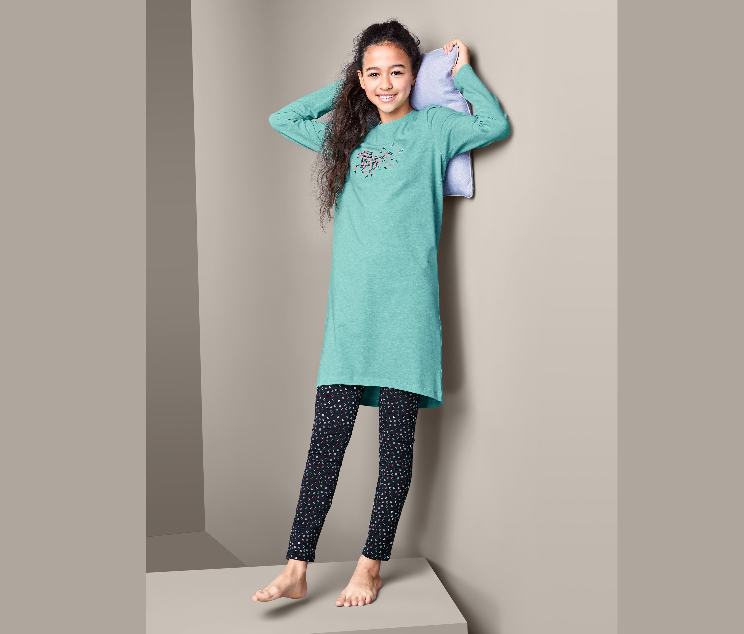 Mädchen mit Kissen hinter dem Kopf steht auf einer grauen Fläche und trägt ein Jersey-Nachthemd mit Leggings.