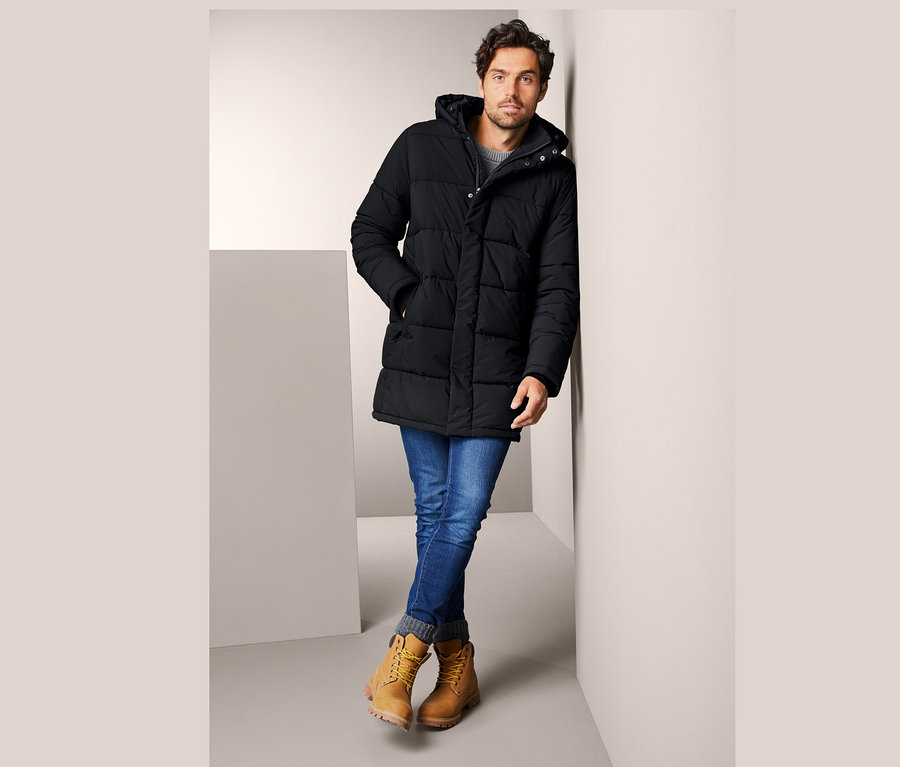 Mann lehnt an Wand, trägt Stepp-Parka mit Kapuze, Jeans und gelbe Stiefel.