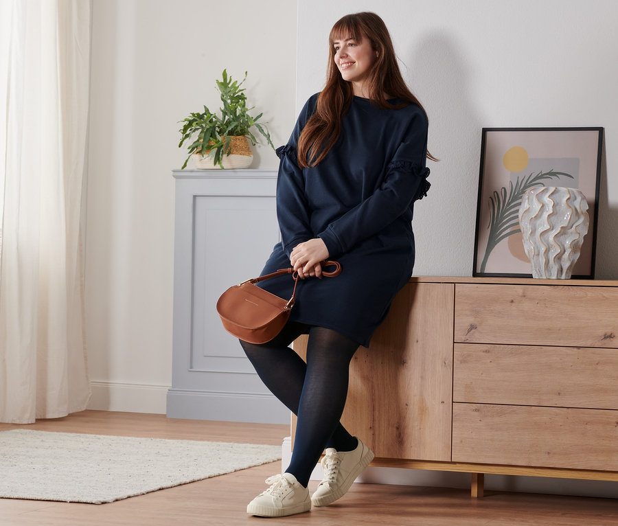 Frau sitzt auf Kommode, trägt Sweatkleid, Leder-Sneaker und Umhängetasche.