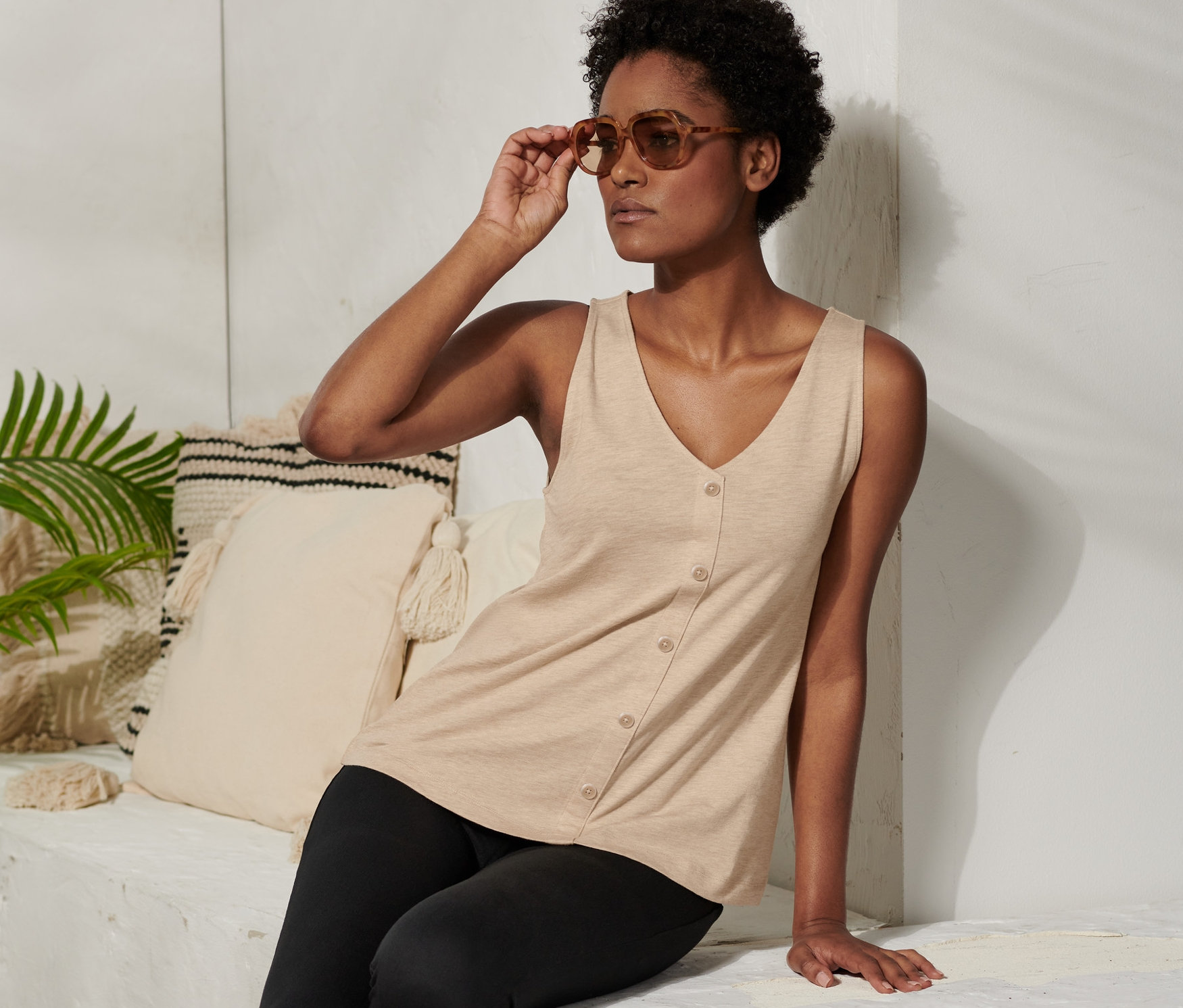 Frau sitzt und justiert ihre Sonnenbrille. Sie trägt ein beiges Top mit Knopfleiste und schwarze Leggings.