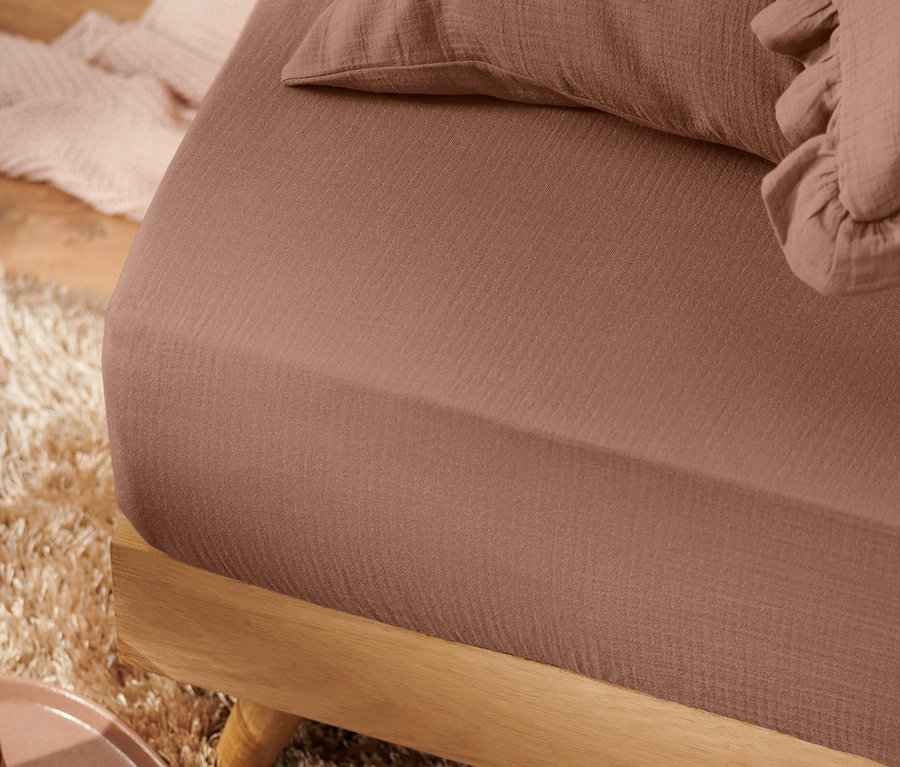 Detailaufnahme eines braunen Premium-Musselin-Spannbettlakens auf einem Bett mit Holzrahmen und braunem Teppich.