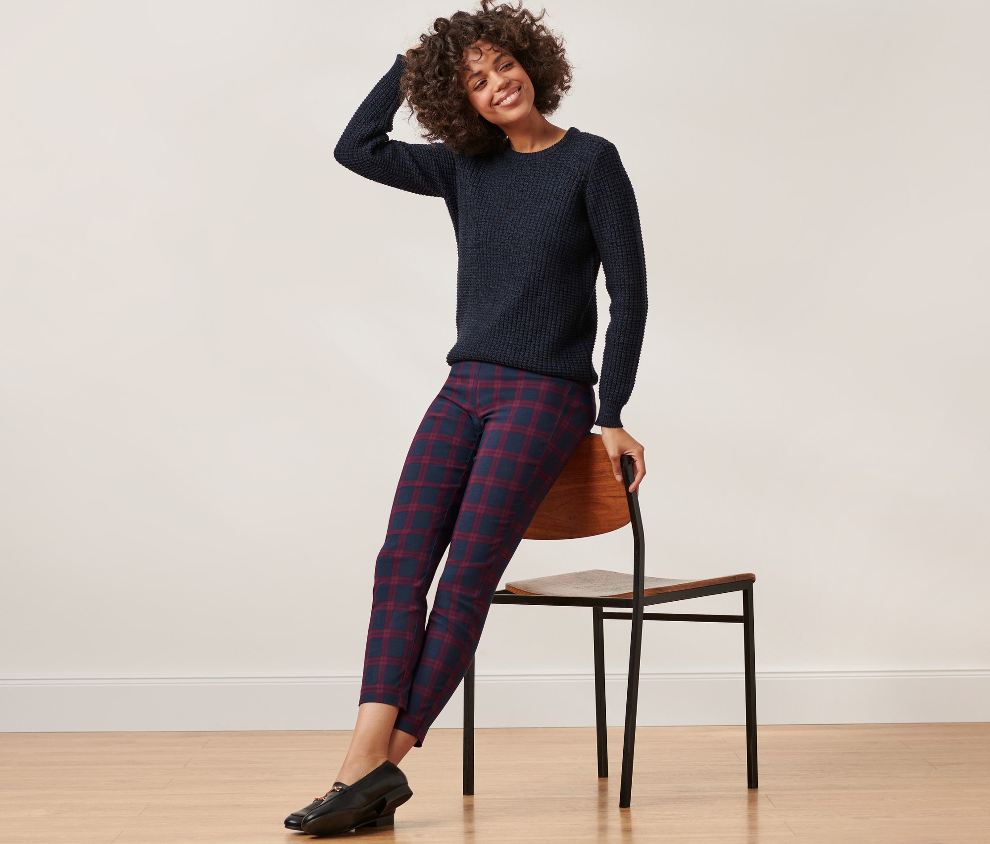 Frau sitzt auf einem Stuhl und trägt einen blauen Pullover und eine rot-blau karierte Stretchhose.