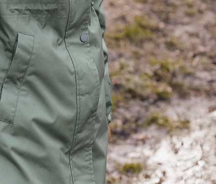 Ein Mädchen steht draußen in einer Blue Denim High-Waist-Jeans, braunen, recycelbaren Gummistiefeln und einem grünen Regenparka.