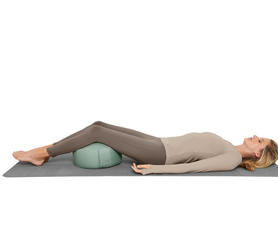 Frau liegt auf einer Matte, mit einem Meditationskissen unter dem Rücken.