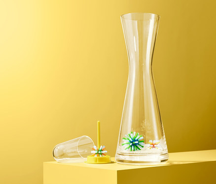 Eine klare Karaffe steht neben einem gelben Halter mit 3 Flaschen-Reinigungsbällen vor gelbem Hintergrund.