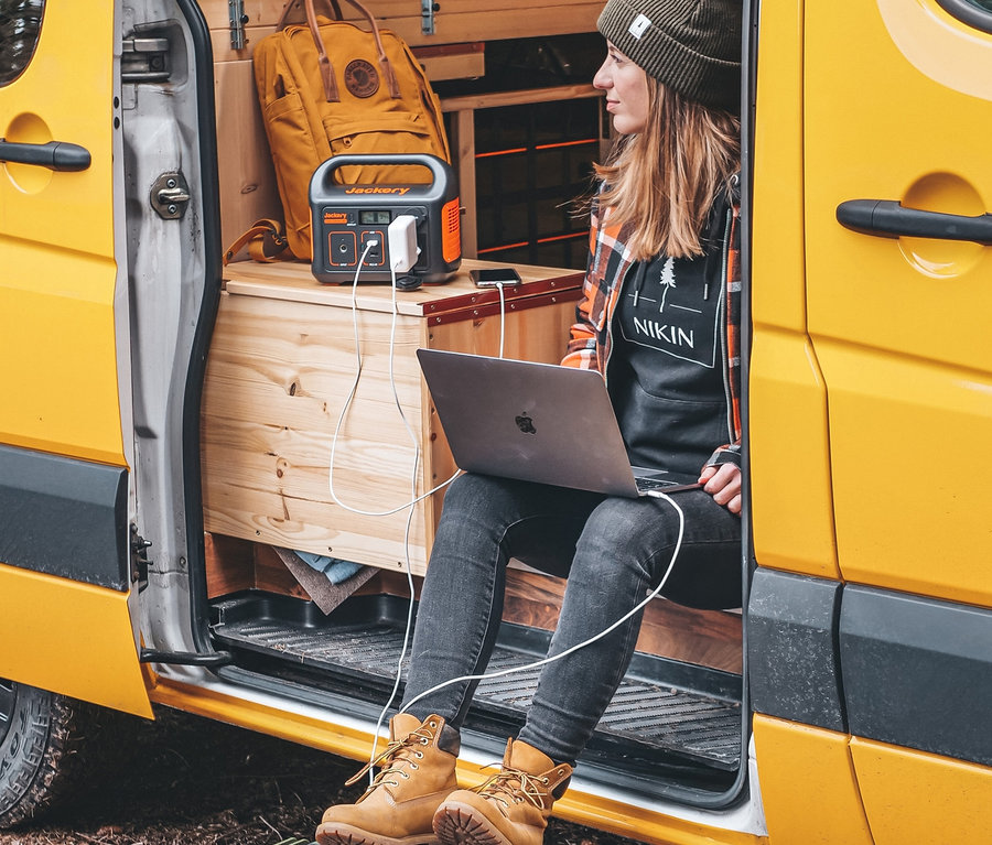 Frau sitzt in einem Lieferwagen und arbeitet an einem Laptop, ihre Füße ruhen auf einem grünen Koffer. Jackery Explorer 240 und SolarSaga 100 sind hinter ihr.