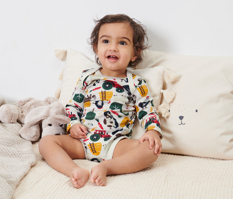 Ein Baby sitzt auf einem Bett und trägt einen 2er-Pack langärmliger Bodys mit Tiermotiven.