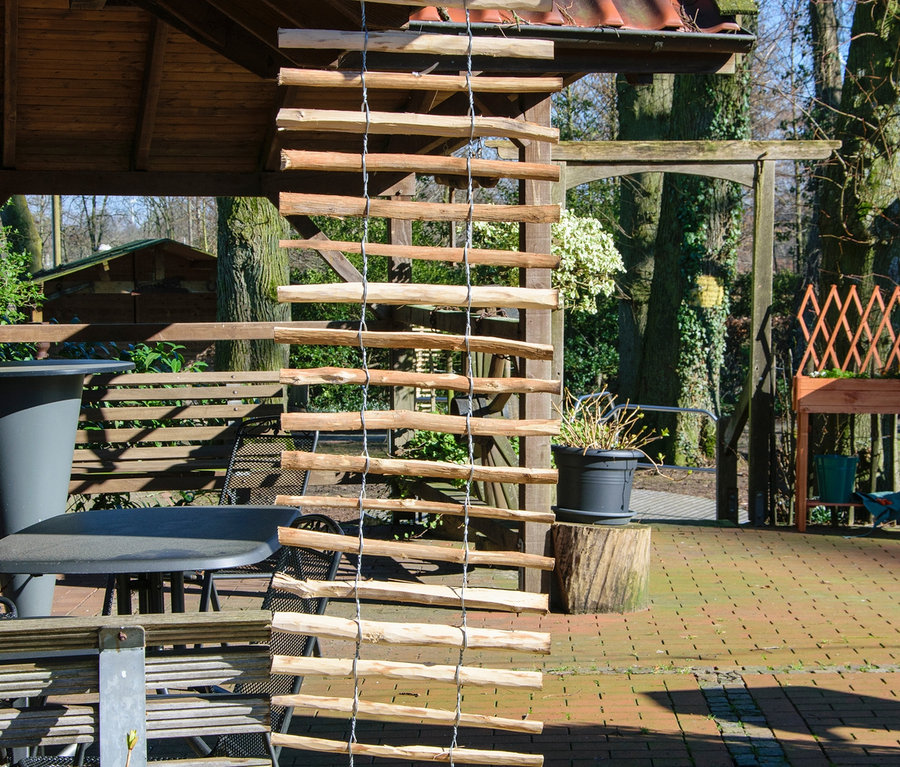 Ein Dobar Rankgitter-Spalier aus Holz hängt von einer Holzkonstruktion auf einer Terrasse.