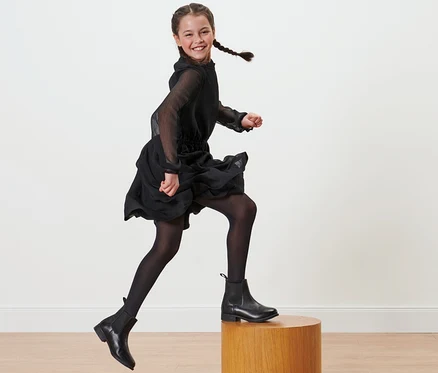 Lächelndes Mädchen mit Zöpfen in schwarzem Kleid und schwarzen 2 Kinder-Nylon-Strumpfhosen springt von einem Holzsockel.