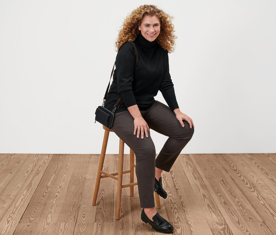 Frau sitzt auf einem Hocker und trägt einen schwarzen Feinstrick-Rollkragenpullover, eine braune Bengalin-Hose, Leder-Loafer und eine Mini-Tasche.