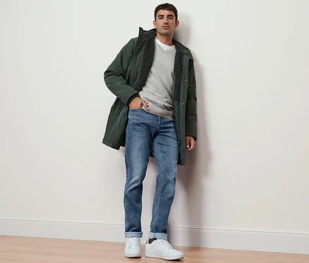 Mann steht an einer Wand, trägt einen grauen Pullover mit V-Ausschnitt, Jeans und eine Parka.