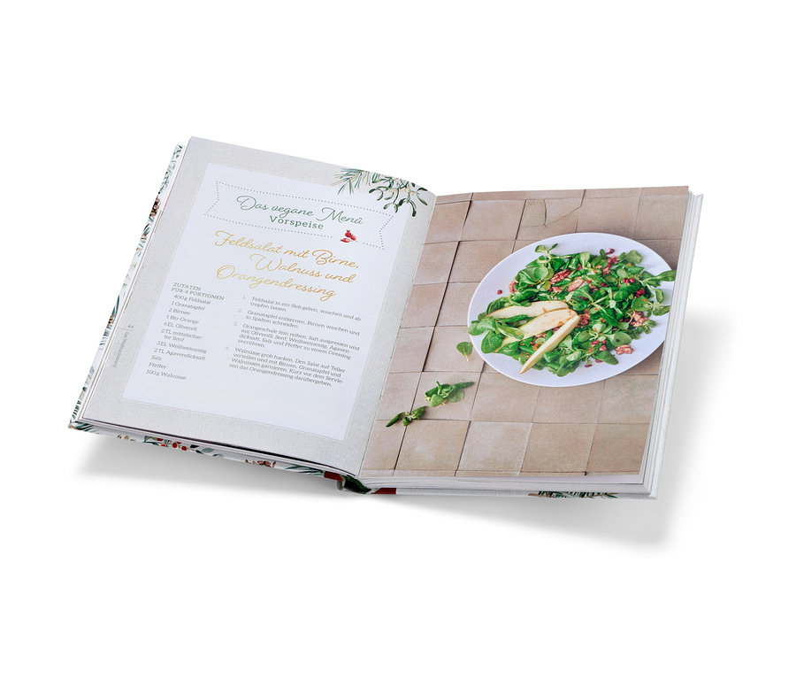 Aufgeschlagenes Kochbuch »Christmas is a feeling« mit Rezept für Feldsalat mit Birne und Walnussdressing und fertigem Salat auf einem Teller.