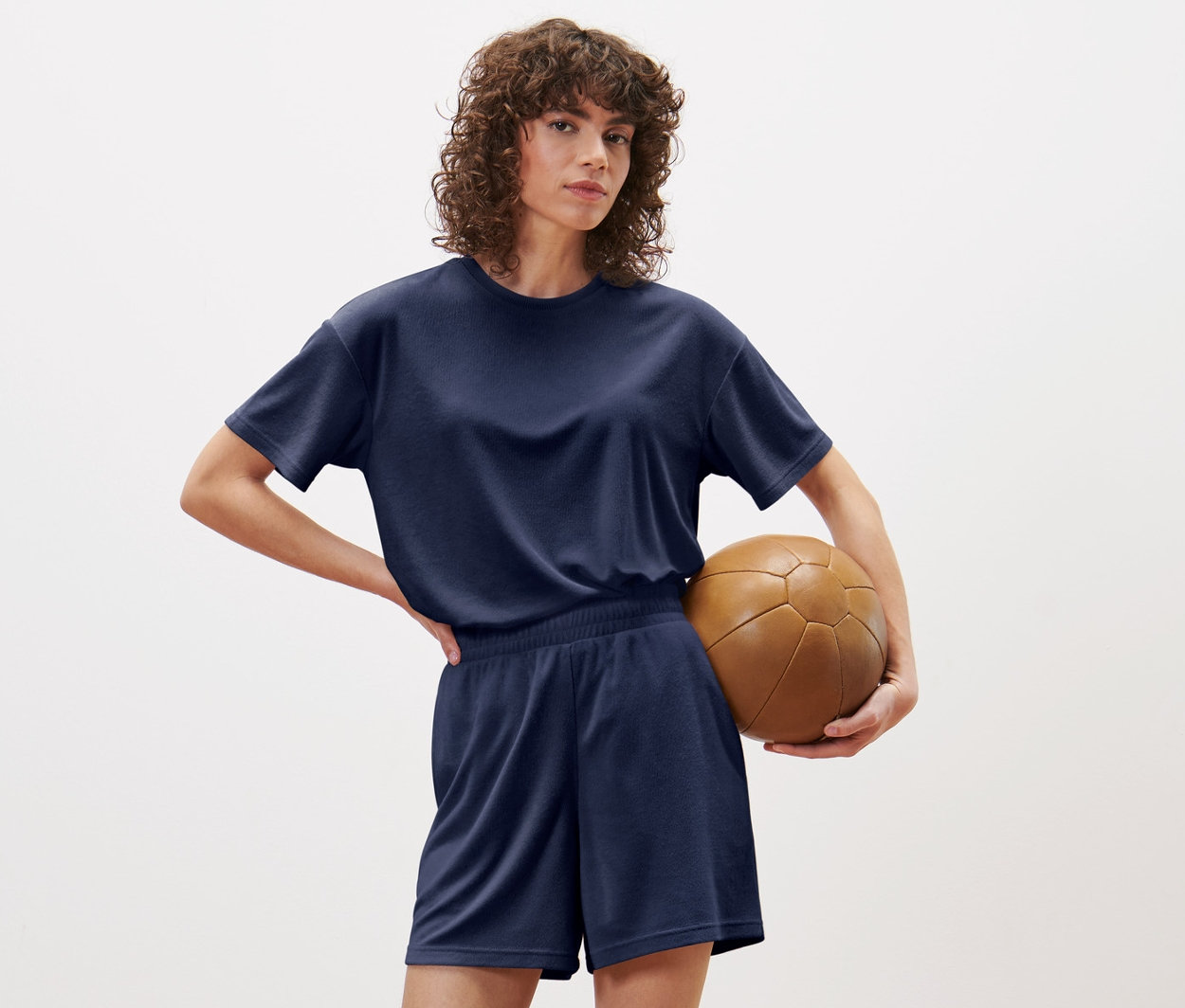 Frau posiert in blauen Frottee-Sportshorts und T-Shirt und hält einen braunen Fußball.