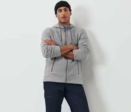 Mann posiert mit verschränkten Armen in einer grauen Strickfleece-Jacke und einer schwarzen Mütze.