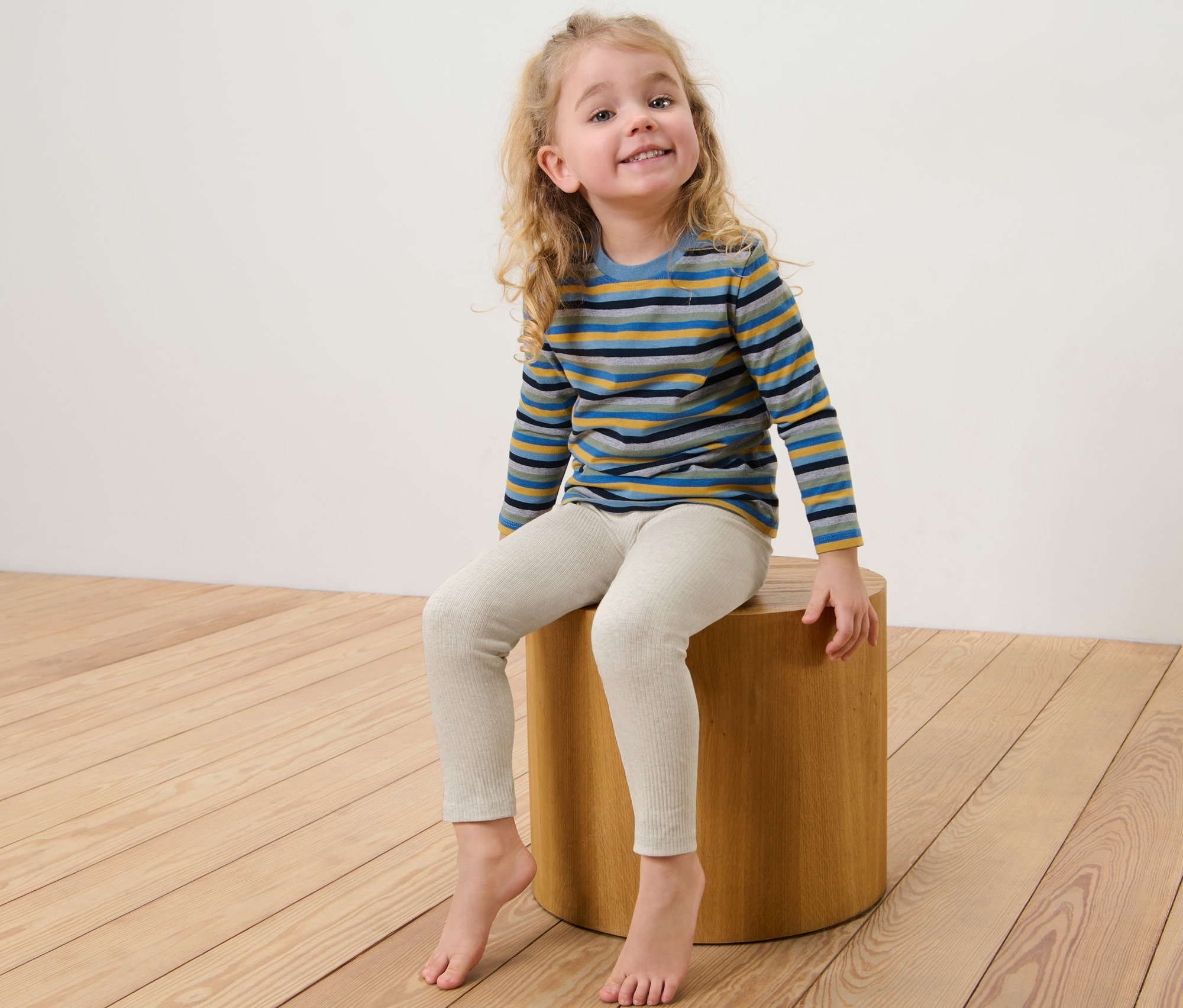 Ein Mädchen sitzt auf einem Holzhocker und trägt 3 Rippleggings und ein gestreiftes Hemd.