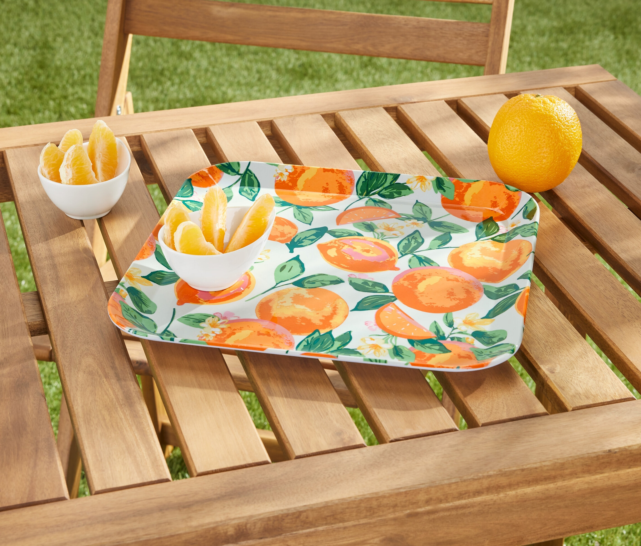 Auf einem Holztisch stehen ein Tablett mit Orangenmuster, eine Schale mit Orangenstücken und eine ganze Orange.