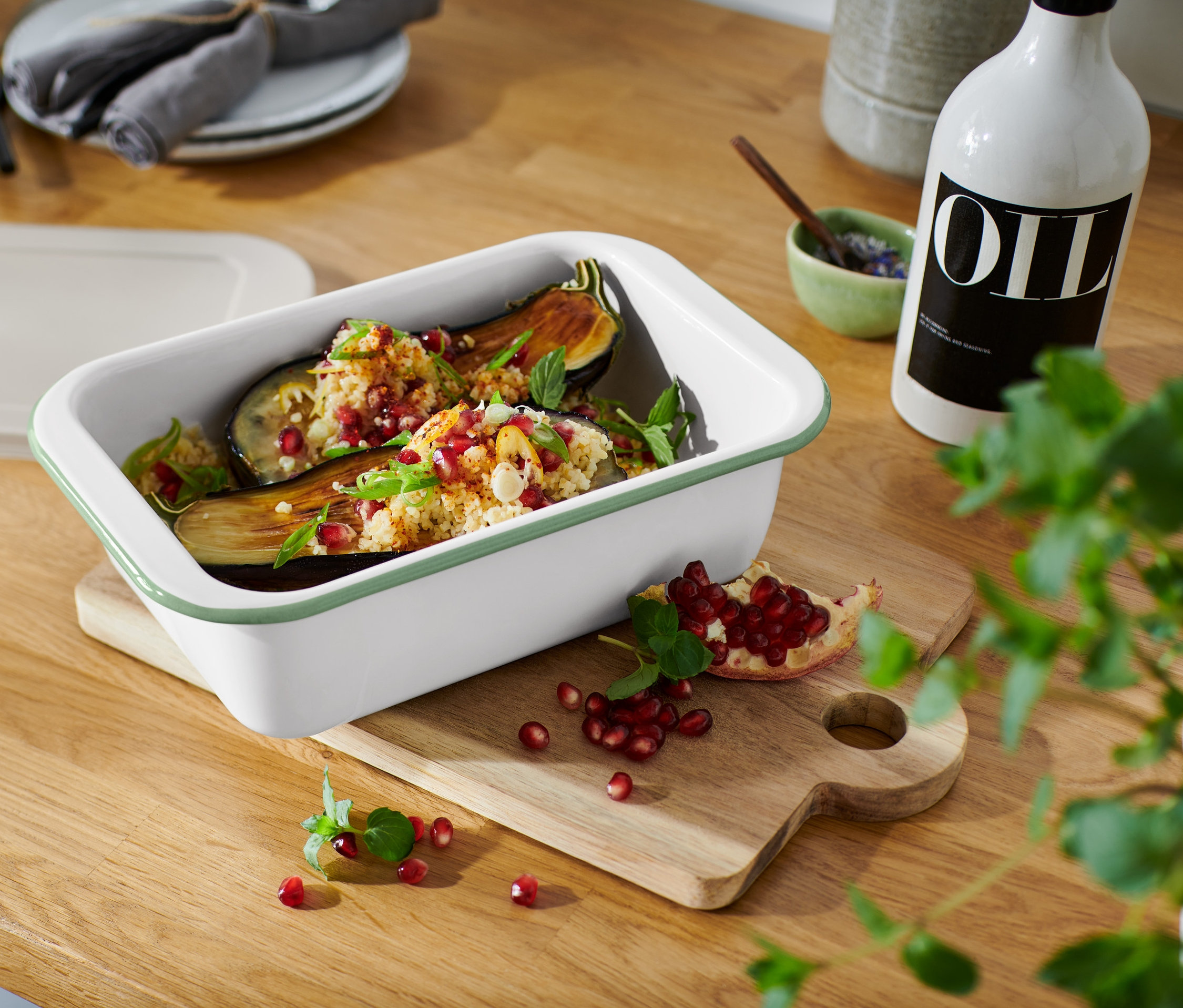 Gebackene Auberginen in einer kleinen Emaille-Auflaufform mit Deckel, mit Granatapfel bestreut, auf einem Holzbrett.