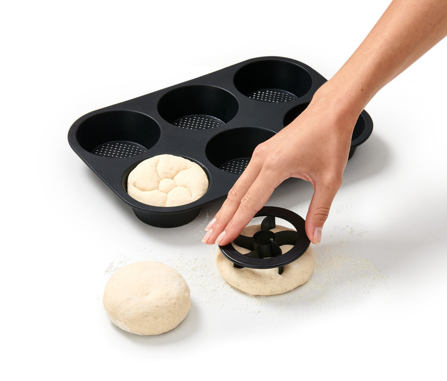 Eine Hand verwendet einen Teigausstecher neben einer perforierten Brötchenbackform.